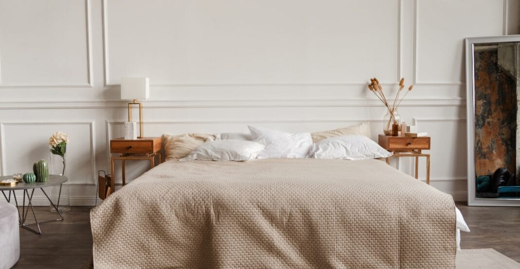 Alt Text: A minimalist bedroom with a neatly made bed covered in a beige textured blanket, white pillows, and two wooden bedside tables, each with a lamp and decorative items. There is a large mirror leaning against the wall on the right, and a small round side table with decorative objects on the left.