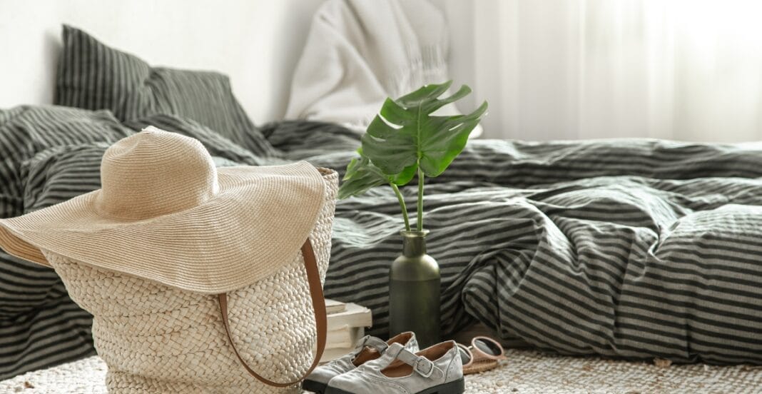 A cozy, well-made bed with dark striped bedding, a large straw sun hat, a green vase with a monstera leaf, a pair of light-colored shoes, sunglasses, and a small stack of books on a textured cream-colored rug in front of sheer white curtains.
