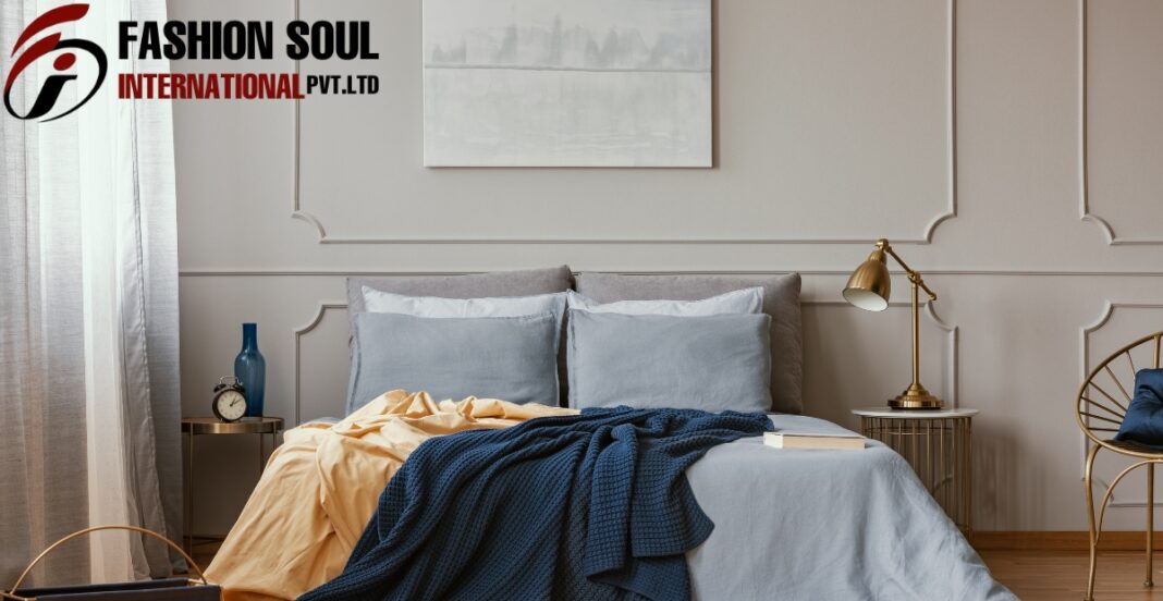 A neatly made bed with gray and light blue pillows and bedding, topped with a dark blue blanket and a yellow sheet, in a modern bedroom with stylish wall paneling. To the left, there is a nightstand with a blue vase and a small clock, while on the right, a gold-colored bedside lamp and a wire chair with a dark pillow are visible.