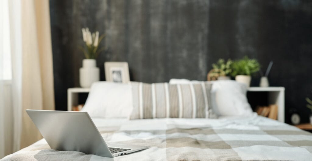 A silver laptop is placed on a bed with a striped beige and white bedspread in a cozy bedroom, with a dark accent wall in the background.