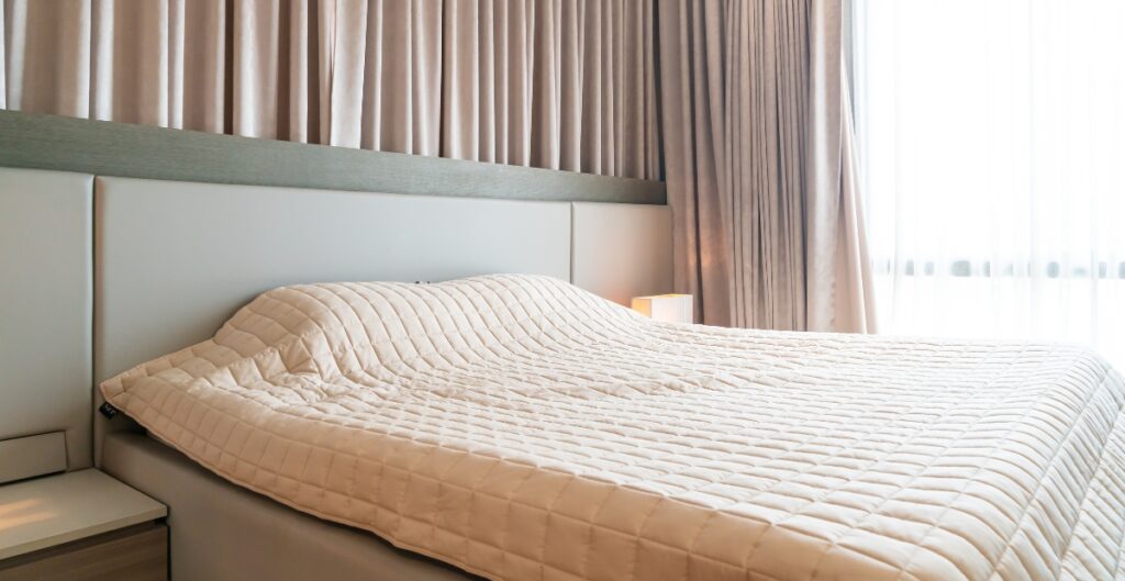 A neatly made hotel bed with a padded beige quilt, situated in a modern, well-lit room with beige curtains and sheer white drapes.