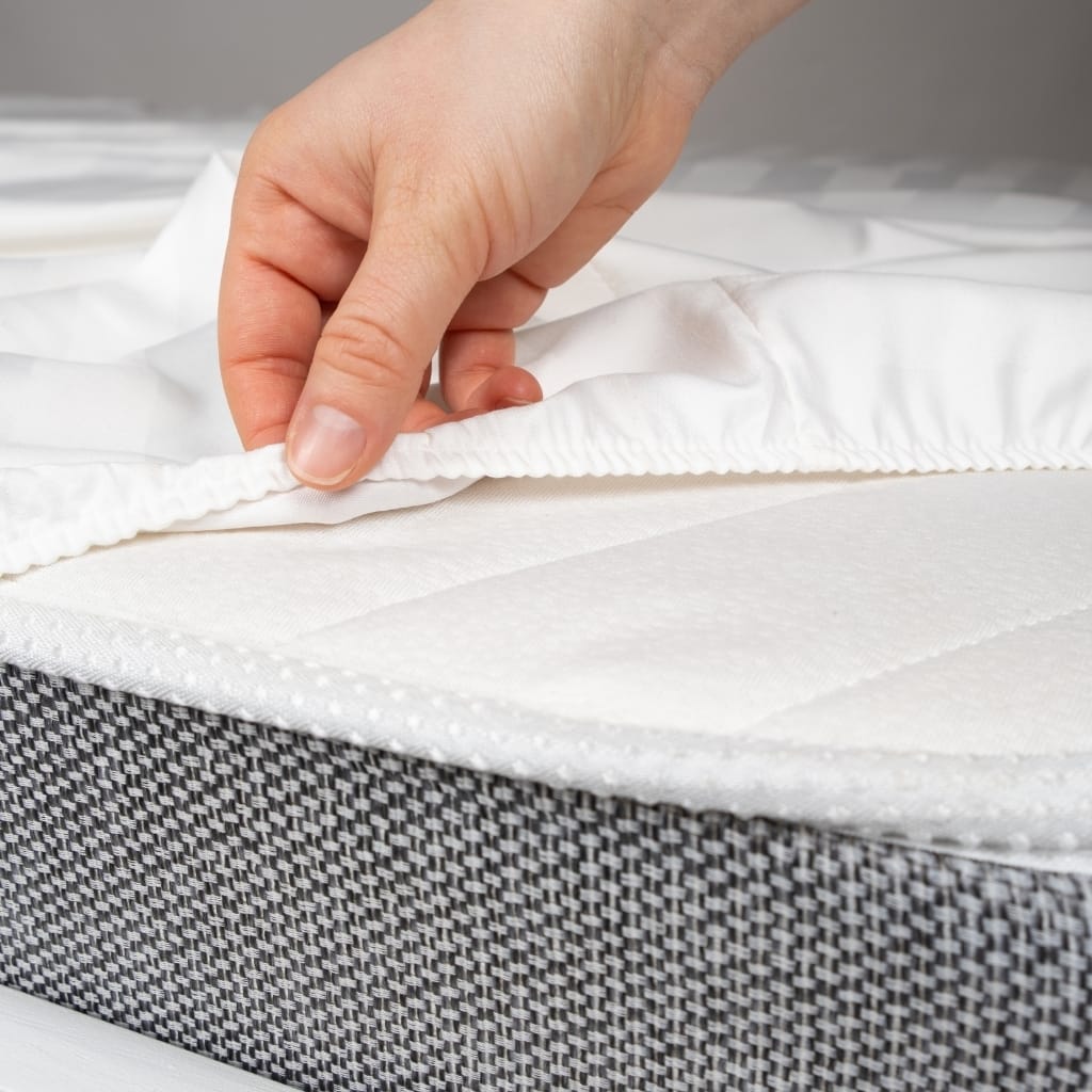 A person's hand is lifting the corner of a white mattress cover or fitted sheet to reveal the mattress beneath.