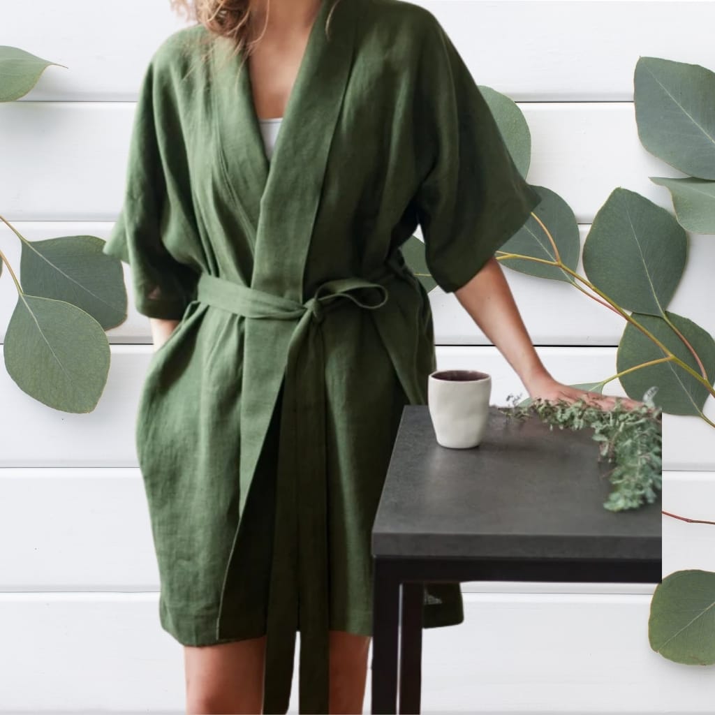 A woman wearing a loose-fitting, olive-green robe-style dress tied at the waist, standing beside a black table with a white ceramic cup and a small plant. Her face and upper body are not visible, and her right arm rests on the table. Large green leaves are arranged on a white wooden wall behind her.