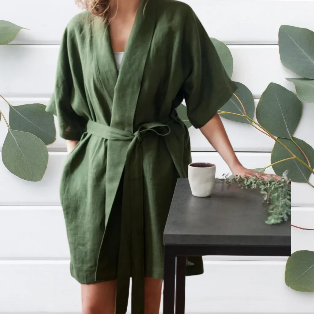 A woman wearing a loose-fitting, olive-green robe-style dress tied at the waist, standing beside a black table with a white ceramic cup and a small plant. Her face and upper body are not visible, and her right arm rests on the table. Large green leaves are arranged on a white wooden wall behind her.