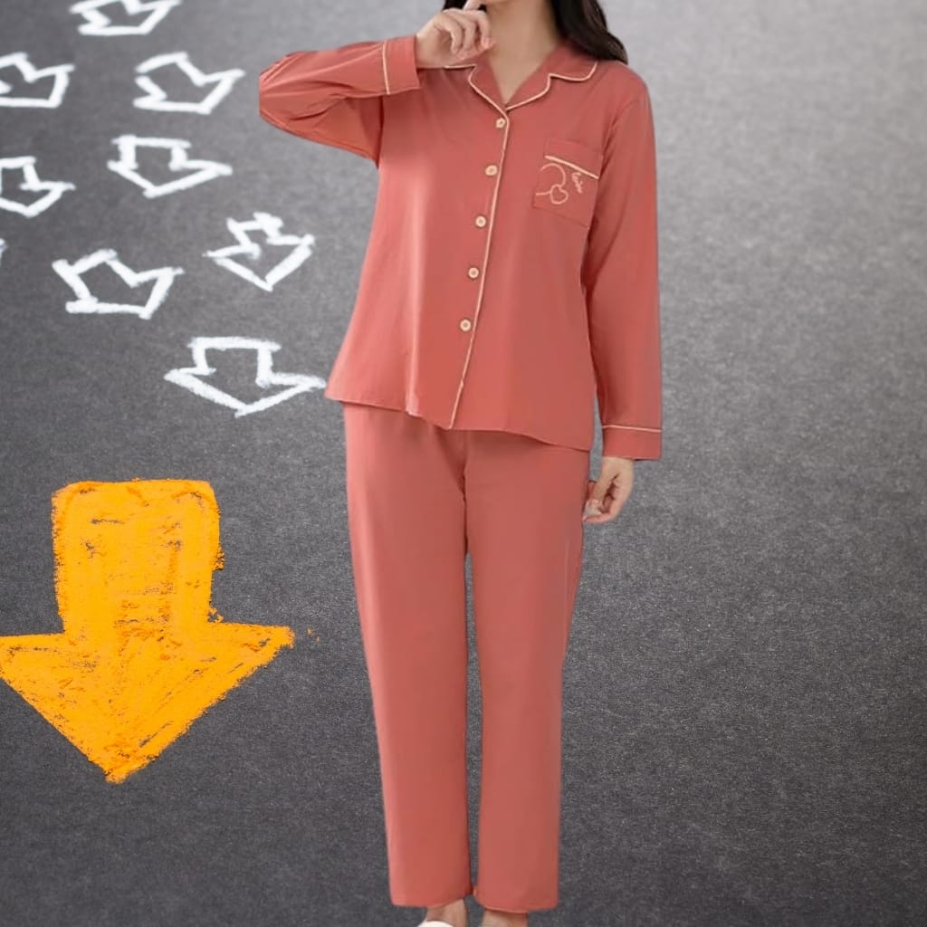 A woman is standing on a textured, dark gray surface with white arrow symbols drawn on it. She is wearing matching coral-colored pajamas with yellow piping details. Her pajama top has buttons and a pocket on the left side, with an embroidered design. She is touching her chin with her right hand, and her face is not visible in the image.