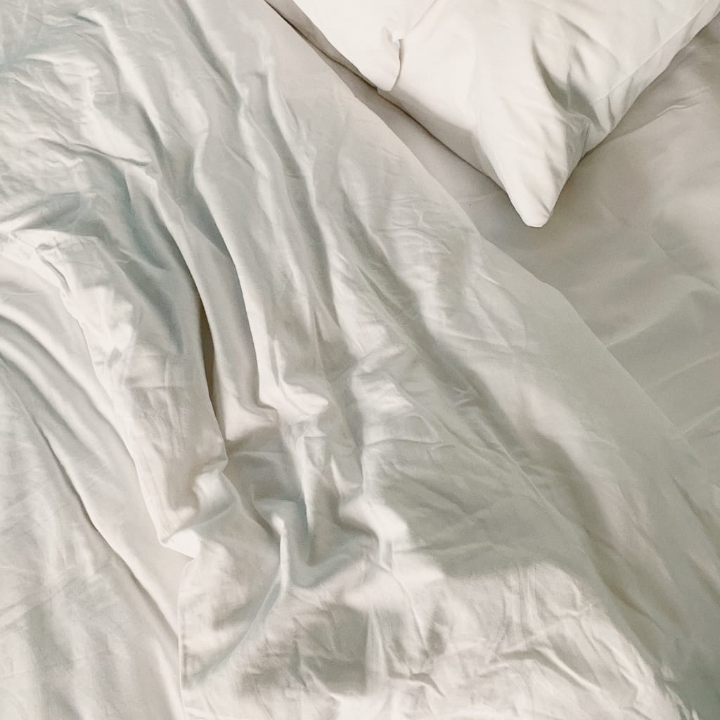 A close-up of wrinkled, cream-colored satin fabric on a bed, with a pillow partially visible in the top right corner.