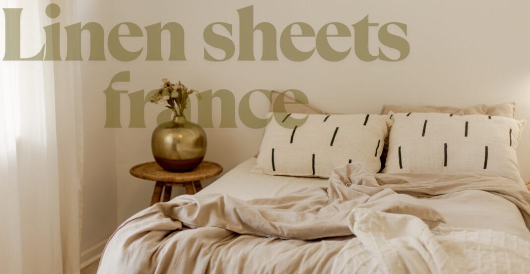 A cozy bedroom with a neatly made bed featuring linen sheets and pillowcases, a small wooden side table with a gold and brown vase holding dried flowers, and sheer white curtains hanging on the left.