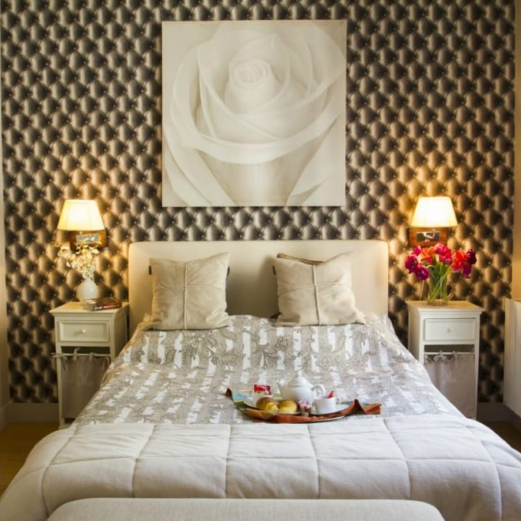 A cozy bedroom with a white bed, two nightstands with lamps, and a large floral artwork on a patterned wall, decorated with flowers and breakfast items on the bed.