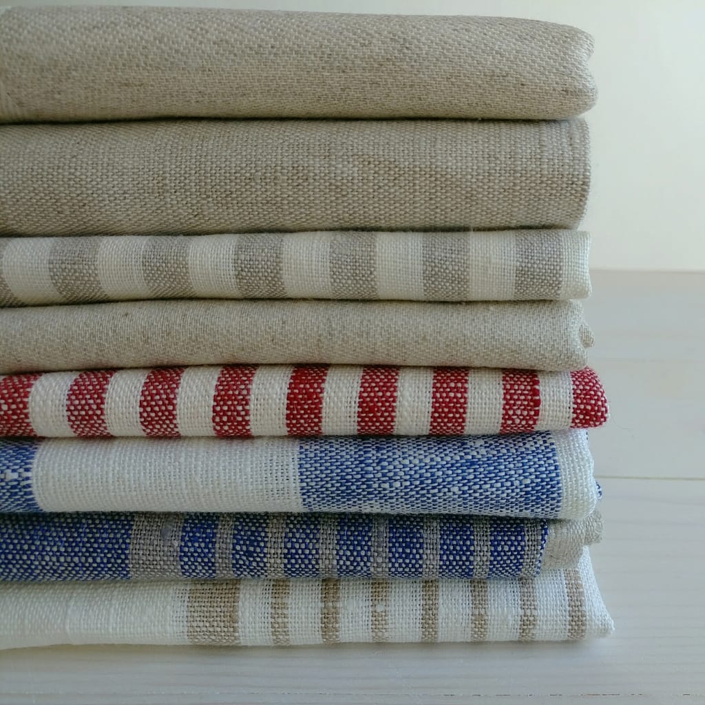 Stack of neatly folded linen fabrics in various shades and patterns, including beige, cream, red, and blue, placed on a white surface.