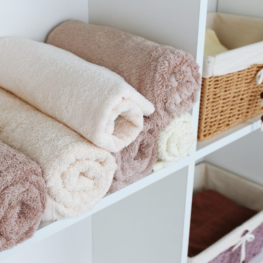 A neatly arranged shelf with soft, rolled towels in neutral tones, and woven baskets for storage, in a bright, clean space.