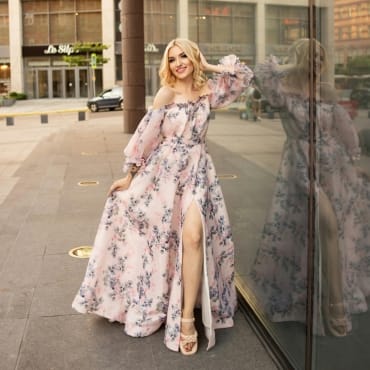 A woman smiling outdoors in a flowing floral maxi polyester dress with a slit, paired with beige sandals, touching a glass window.