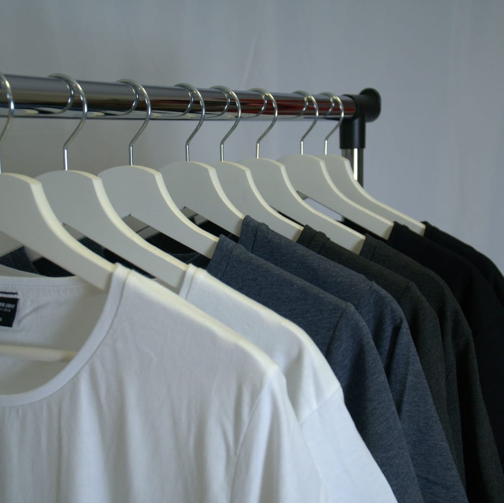 A row of white, gray, and black cotton t-shirts hanging on white hangers on a clothing rack against a neutral background, highlighting summer moisture-absorbing apparel.