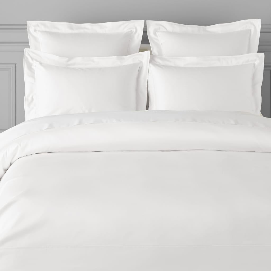 A close-up view of a neatly made bed with white linens, four large pillows, and a wooden headboard, set against a light gray wall with subtle molding.