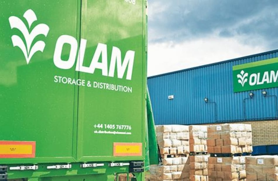 A green Olam Storage & Distribution truck parked outside a warehouse with stacks of bricks, with the Olam logo and contact information visible. Top Global Companies