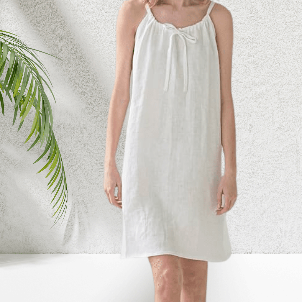 A woman wearing a sleeveless, knee-length white dress with a bow detail at the neckline, standing against a textured white wall with a green palm leaf partially visible on the left.