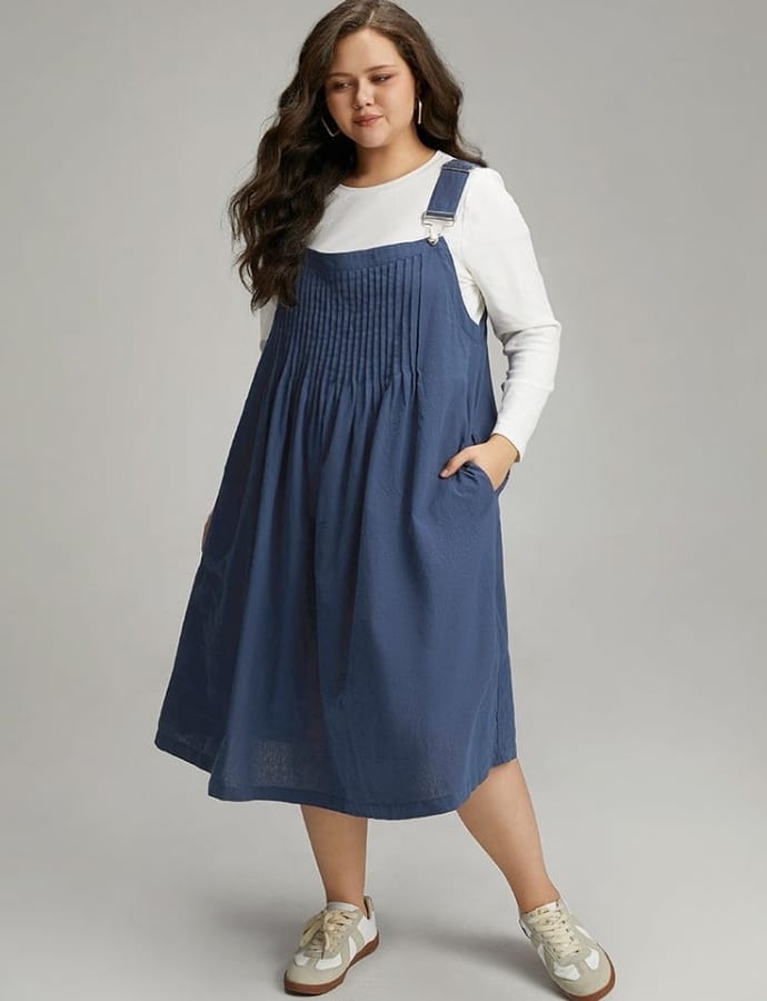 A plus-size woman with long, wavy brown hair wearing a casual denim pinafore dress over a white long-sleeve top, paired with beige sneakers, standing with one hand in her pocket against a neutral background.