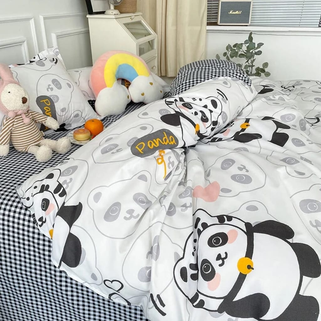A cozy bed with panda-themed bedding, featuring black and white pandas and small yellow and peach accents. The bed is decorated with plush toys, including a stuffed teddy bear in a striped outfit, a rainbow-shaped plush, and a cute panda plush. A small table nearby holds an orange, a peach, and a plate. The background includes a white cabinet, a plant, a window with blinds, and a vintage radio.