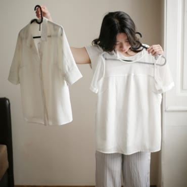 A woman holding up two white polyester shirts, one in each hand, indoors near a window. She is wearing striped pajamas and looking at the shirts with interest.