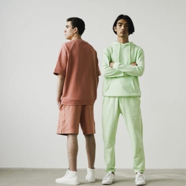 Two young men standing against a plain background, showcasing casual streetwear outfits made from polyester jersey fabric, one in muted pink and the other in light green.