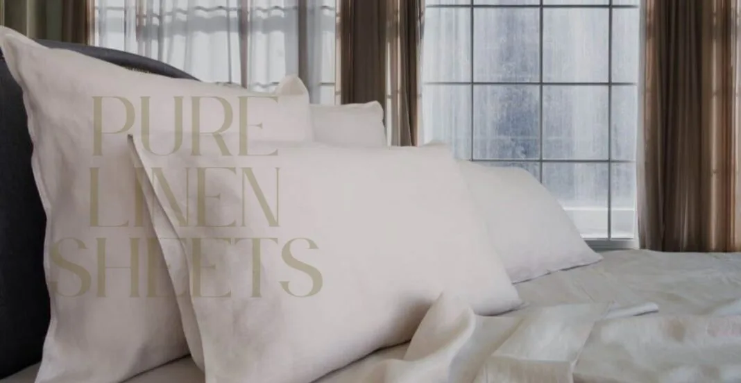 A cozy bed with neatly arranged white linen sheets and matching pillowcases, set in front of large windows with sheer curtains, allowing natural light to fill the room.