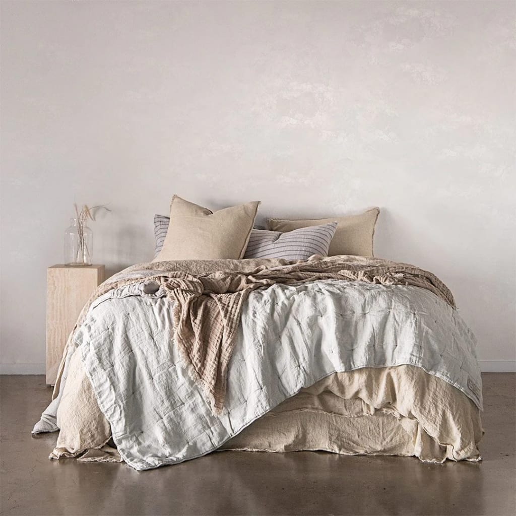 A minimalist, cozy bedroom with a neatly made bed featuring neutral-toned bedding, pillows, and a beige blanket. A small wooden nightstand beside the bed holds a glass vase with dried plants, set against a plain, white wall.