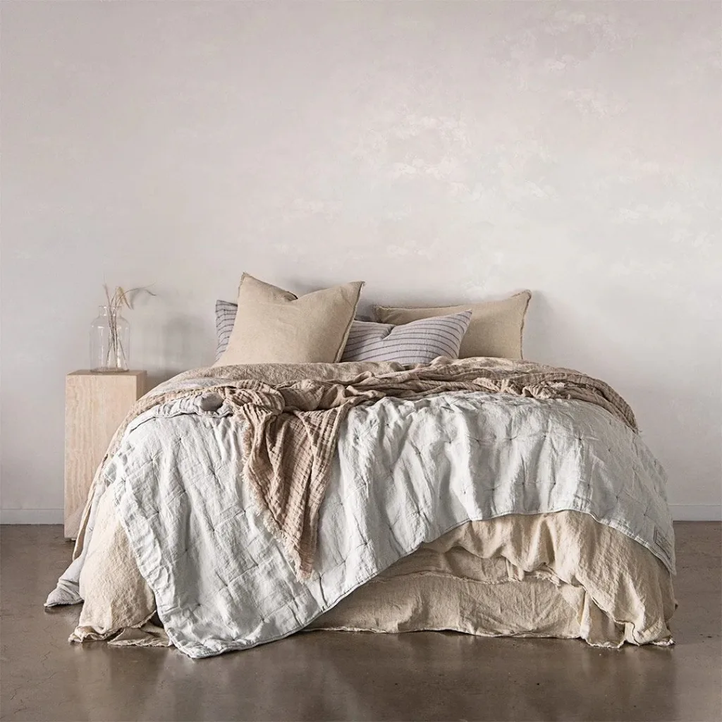 A minimalist, cozy bedroom with a neatly made bed featuring neutral-toned bedding, pillows, and a beige blanket. A small wooden nightstand beside the bed holds a glass vase with dried plants, set against a plain, white wall.