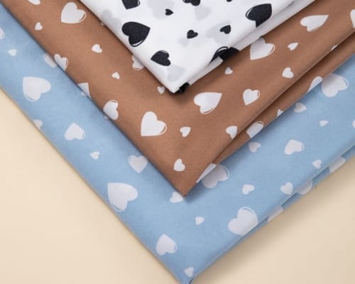 Four pieces of fabric with heart patterns in white, black, brown, and blue, arranged diagonally on a neutral background.