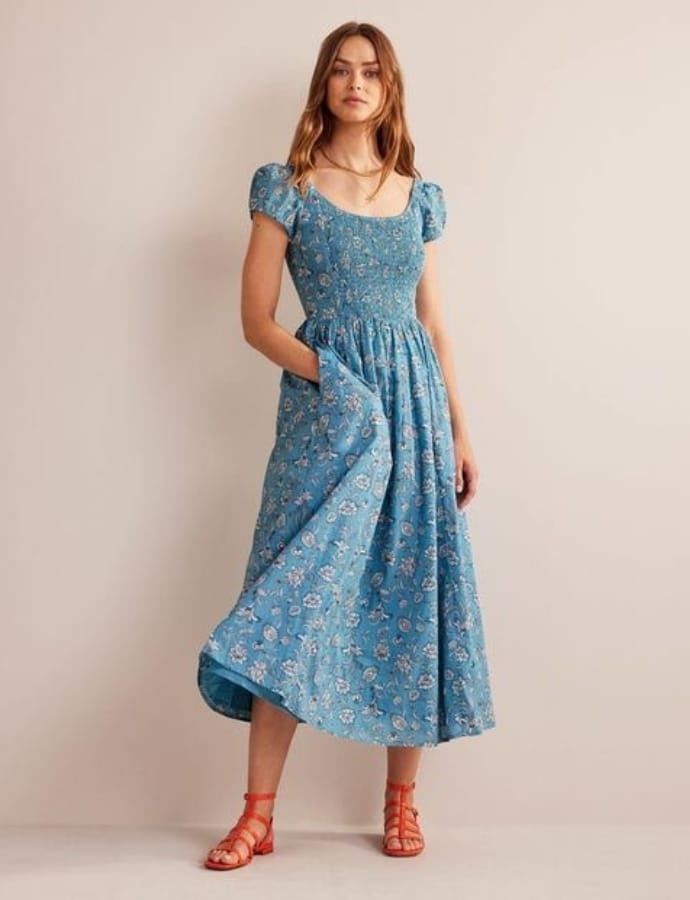 A woman wearing a blue floral smock dress with puffed short sleeves and a gathered waist, paired with red sandals, standing against a plain background.