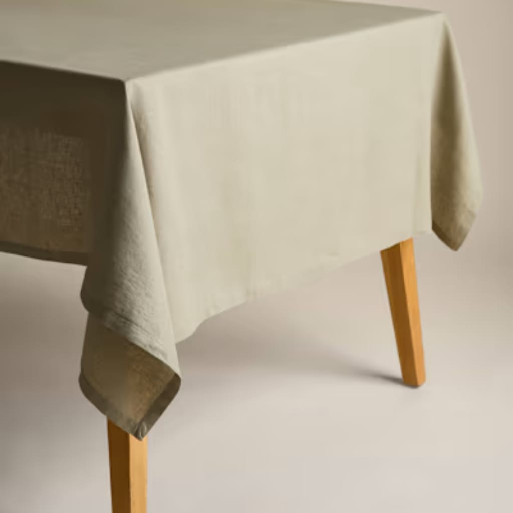 A close-up view of a table covered with an off-white or cream-colored tablecloth that drapes over the edges, with one of the table's legs visible, made of light-colored wood, against a plain neutral background.