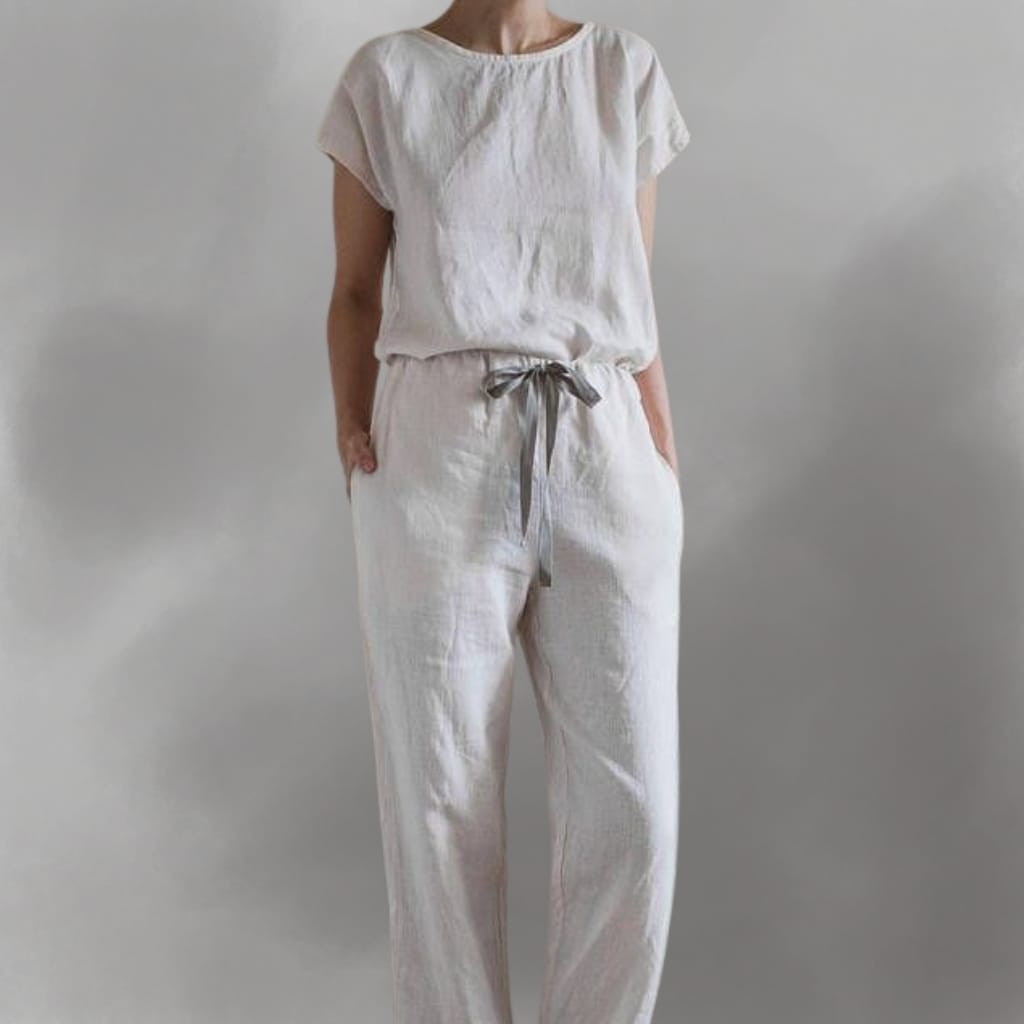 A woman dressed in a white, loose-fitting short-sleeved top and matching white pants with a gray ribbon belt, standing against a plain light gray background.