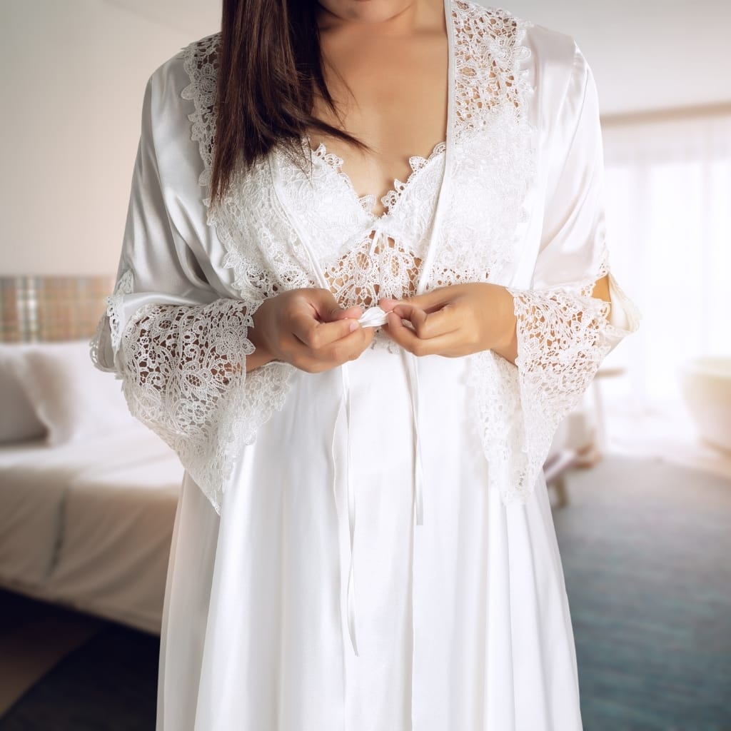A woman wearing a satin and lace nightgown, adjusting the front tie while standing in a softly lit bedroom.