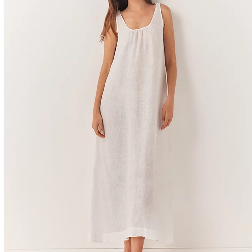 A woman wearing a long, sleeveless, light-colored summer dress with a round neckline, standing against a plain wall.