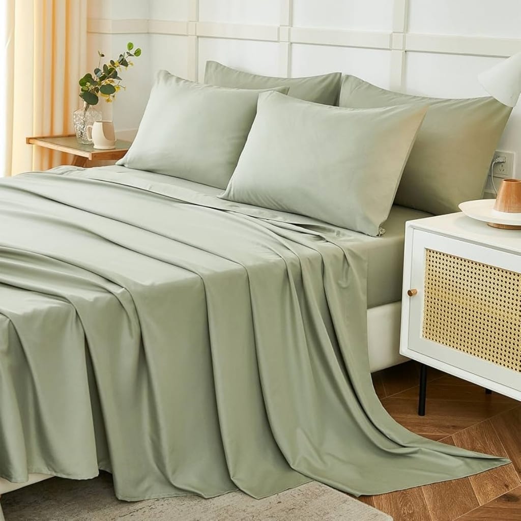 A neatly made bed with light green, silky bedding and matching pillows, set in a bright, modern bedroom with a side table holding a decorative vase.