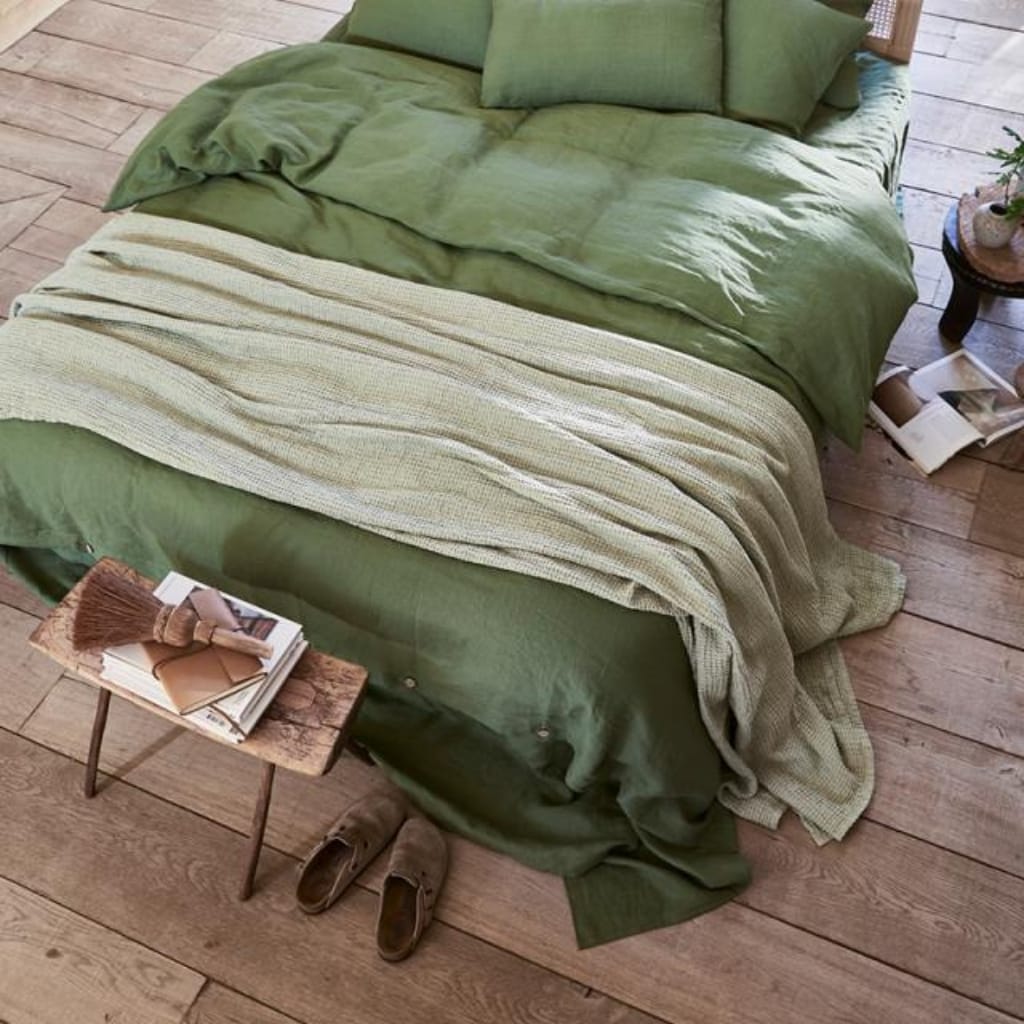 A cozy bed with green bedding and pillows, partially covered with a light textured blanket. There is a small wooden side table next to the bed with an open book and a decorative broom, and a pair of slippers on the wooden floor.