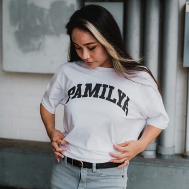 A woman looking down while adjusting her white T-shirt with bold "FAMILY" text, paired with light-colored palazzo pants, standing indoors.