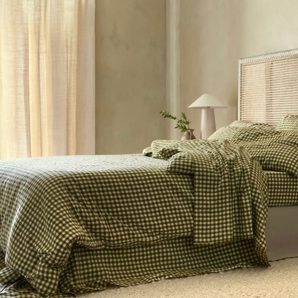 A cozy bedroom featuring a bed with a checkered bedspread in shades of brown and white, against a wall with soft, natural lighting coming through beige curtains. A bedside table holds a white lamp and a small vase with green foliage.