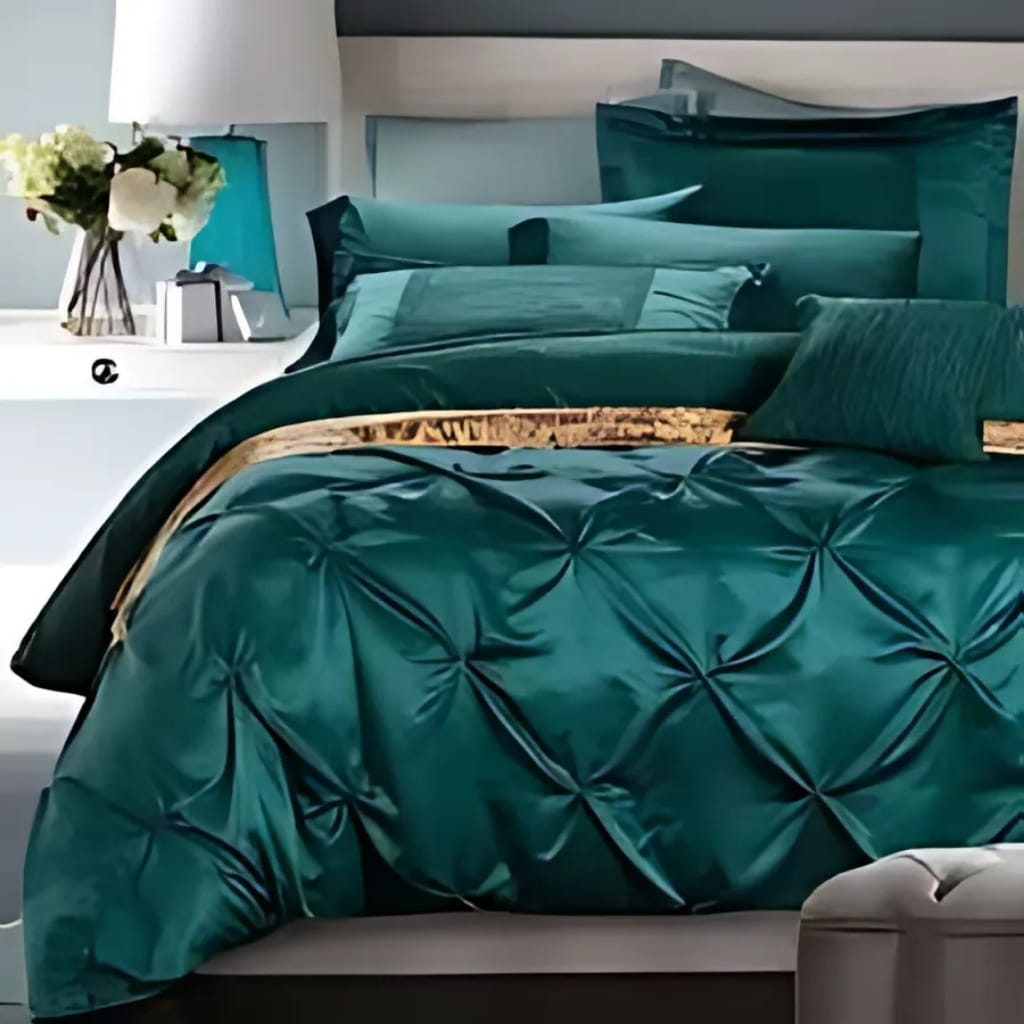 A neatly made bed with emerald green satin bedding, featuring a tufted comforter and matching pillows. There is a white nightstand beside the bed with a contemporary lamp, a small vase of white flowers, and a decorative object. The room has a modern and elegant decor style.
