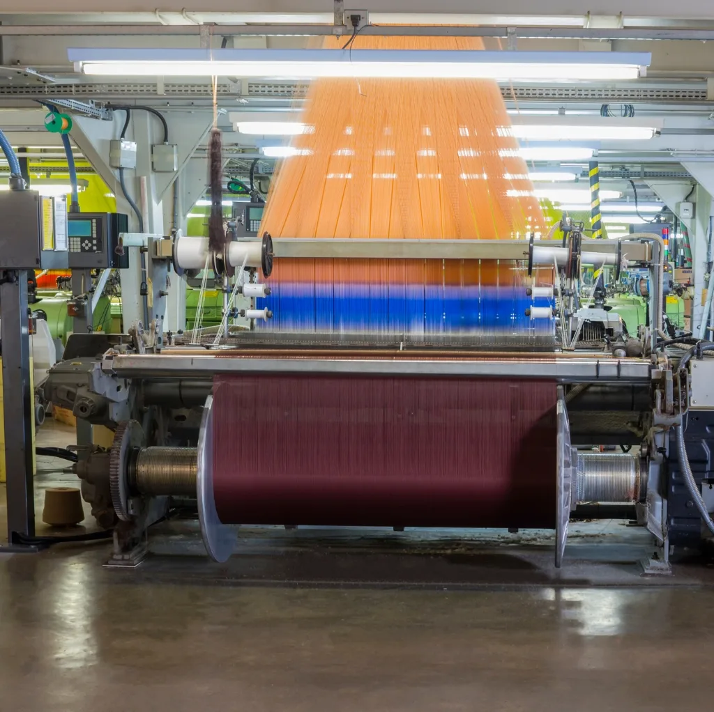 A modern textile manufacturing machine processing brightly colored threads in a factory setting, with vibrant orange, blue, and maroon yarns visible. Cotton Textiles Made in USA