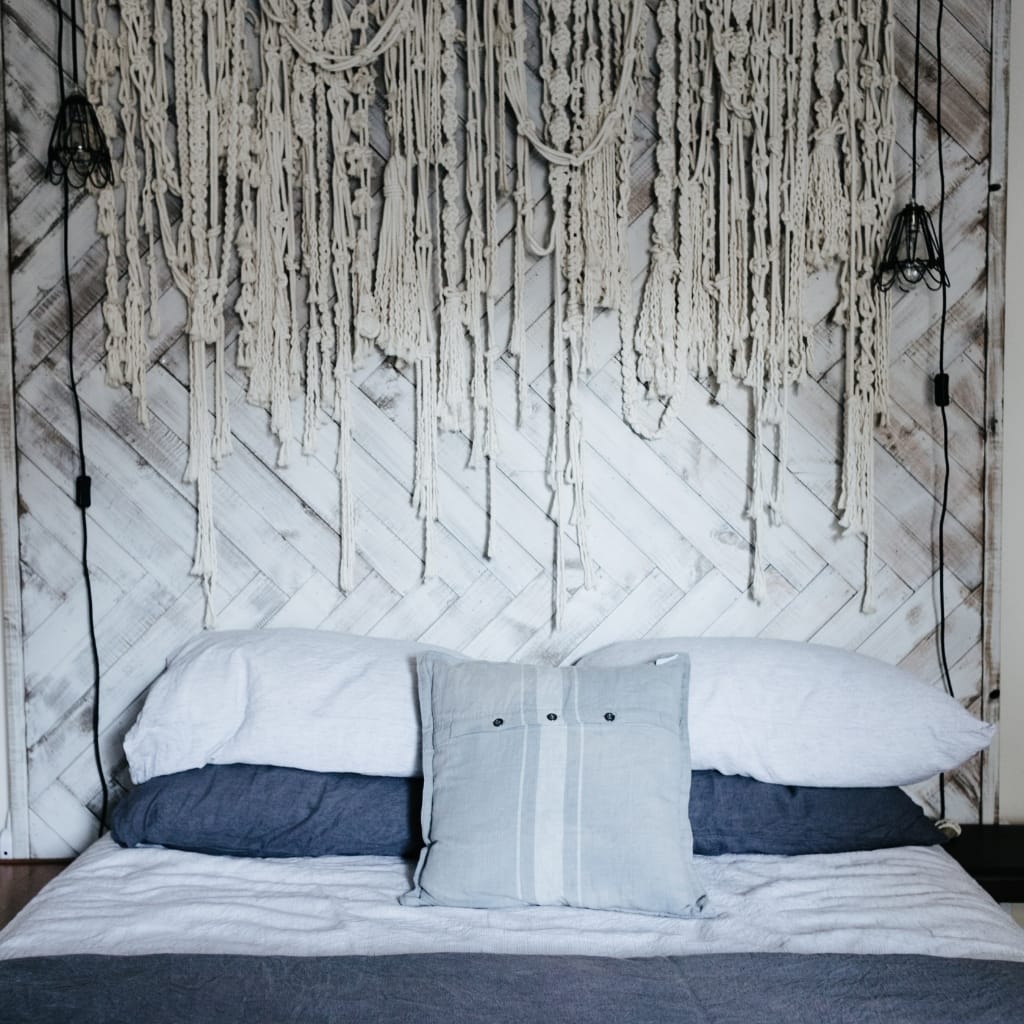 A cozy bedroom scene featuring a bed with white and dark gray bedding and a decorative pillow. The background has a wooden, diagonally paneled wall with a large, intricate macramé wall hanging. Two black wall-mounted lamps with exposed bulbs are on either side of the macramé décor