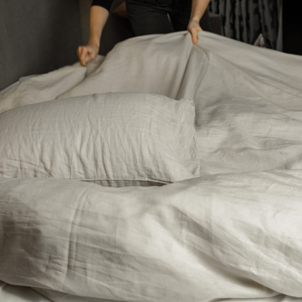 A person is in the process of making a bed, pulling up or arranging a white or off-white bed sheet over a mattress, with a pillow visible in the foreground.