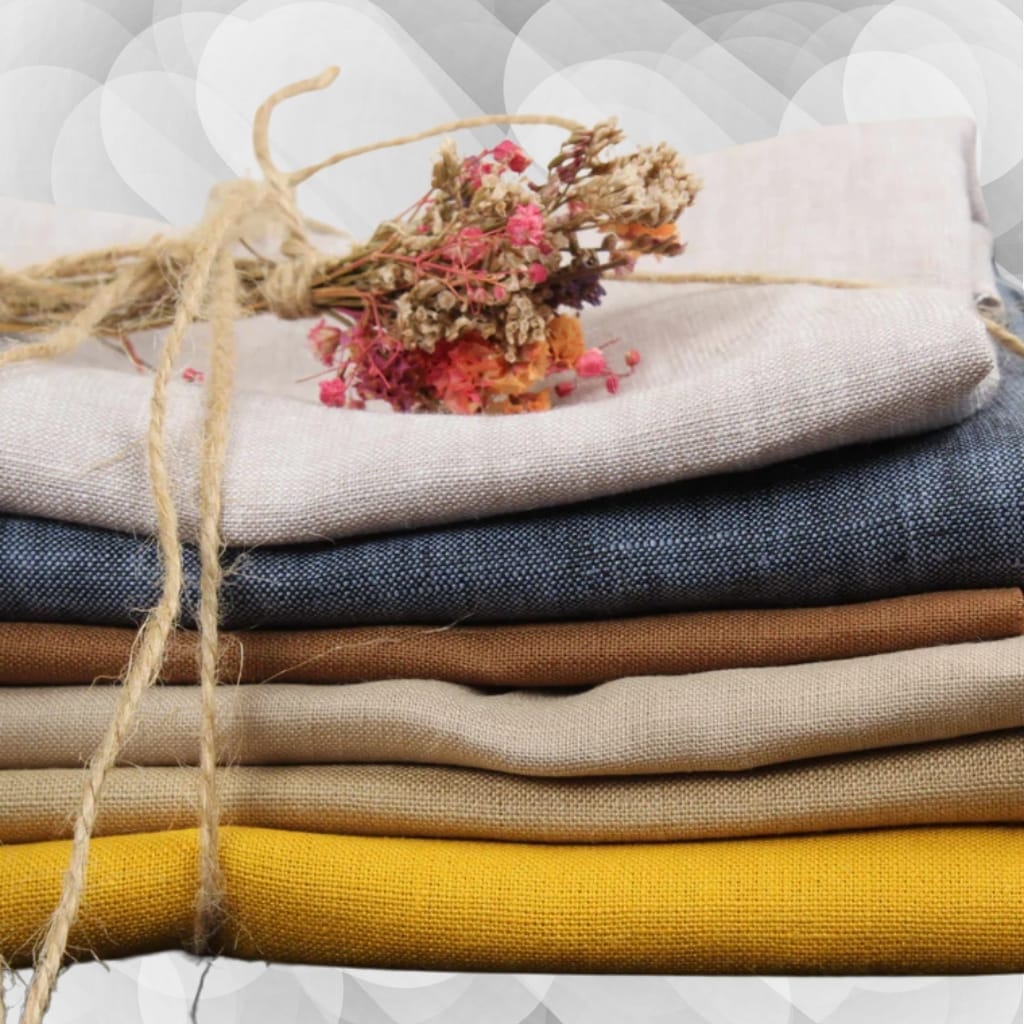 A neatly folded stack of fabric in various colors—light beige, dark gray, brown, another beige, and yellow—tied together with a rustic string that also holds a small bunch of dried pink and beige flowers.