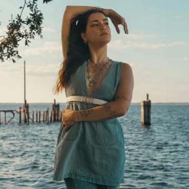 A woman standing by the water with her arm raised, wearing a midi polyester dress with a belt, enjoying a serene outdoor scene.