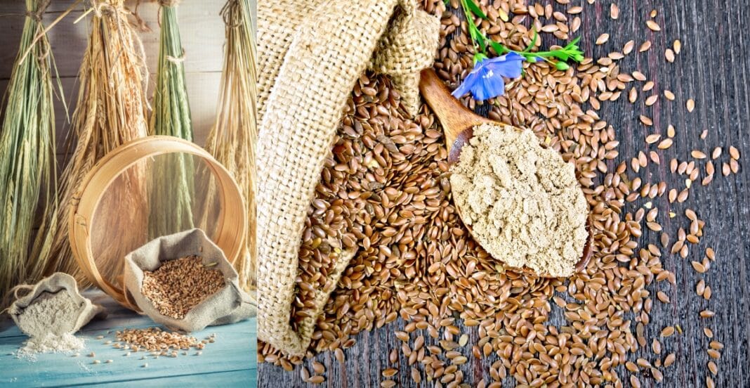 A close-up of flax seeds scattered on a dark wooden surface with a scoop of ground flaxseed, a blue flower, and a small burlap cloth. The background features a basket and bundles of dried plants hanging.