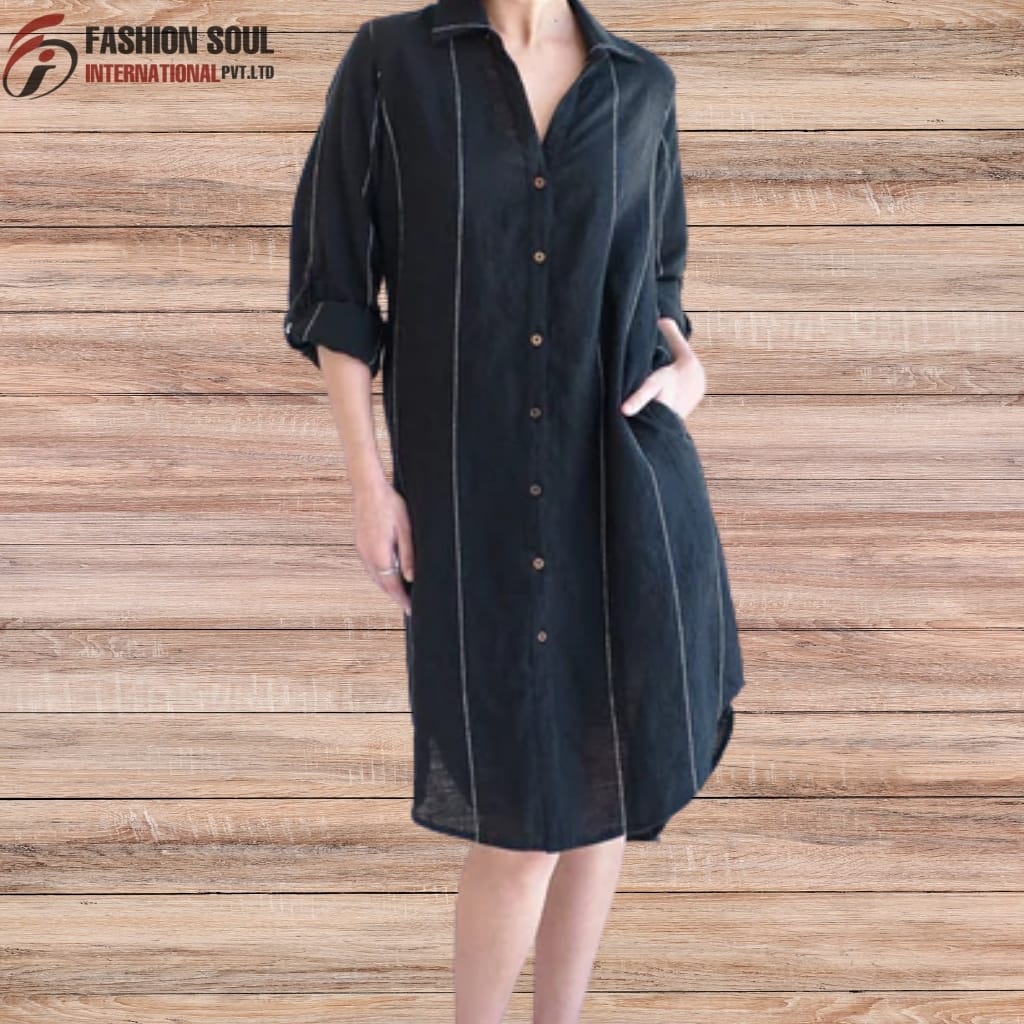 A woman is standing against a wooden background, modeling a long black shirt dress with contrasting vertical white stitches and wooden buttons down the front. She has her right hand in her pocket and her left arm relaxed at her side.
