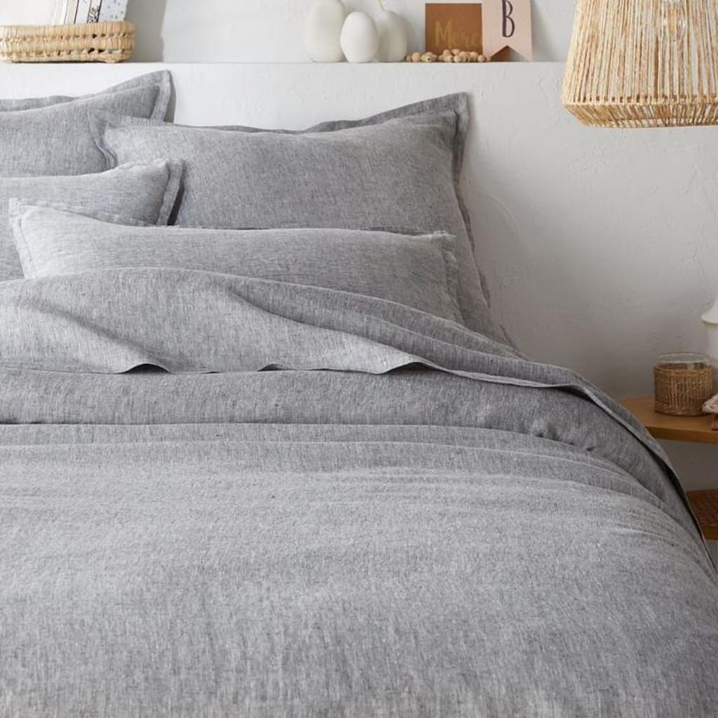 A neatly made bed with light gray bedding and matching pillows, positioned next to a wooden nightstand with decorative items. The wall behind features a shelf with small vases and framed photographs, adding to a cozy, minimalist bedroom decor.