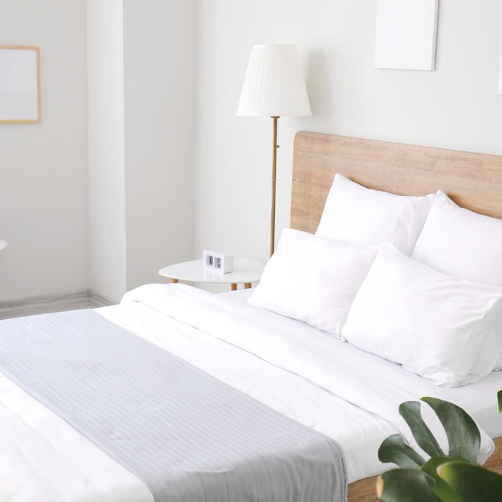 A minimalist bedroom featuring a bed with white linens and pillows, a wooden headboard, a white bedside table with a digital clock, a floor lamp, and a leafy plant in the corner.