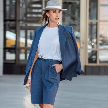 A woman in a stylish outfit, tying a polyester blouse into high-waisted shorts, standing outdoors in an urban setting.