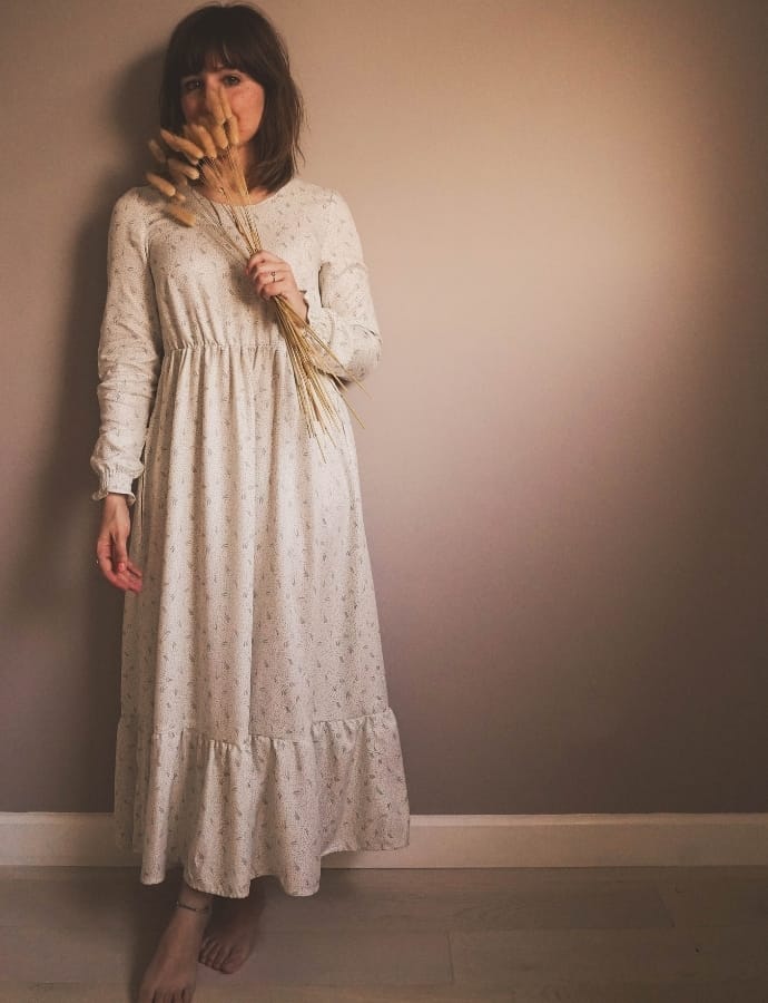 A woman standing against a plain wall, wearing a long, white, tiered cotton dress with floral patterns, holding a small bundle of dried wheat, with her face partially obscured by her hand and her hair in a tousled style.