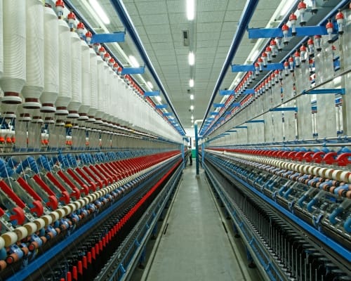 : Rows of spinning machines in a textile factory, with threads and yarns producing cotton fabrics under bright, industrial lighting. Top Cotton Companies