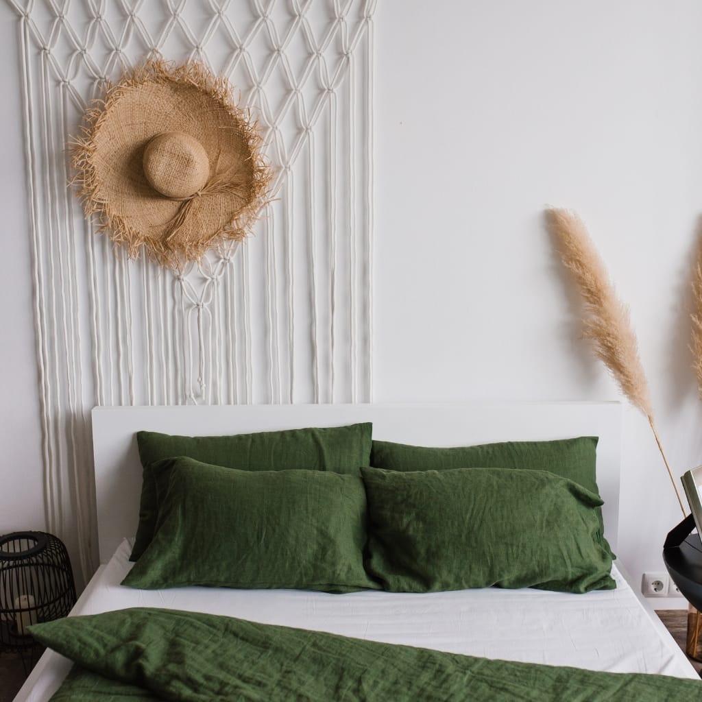 A neatly made bed with dark green velvet pillows and a matching blanket on a white bed frame. Behind the bed, there is a textured white wall with a decorative round straw sun hat hanging above, and soft beige pampas grass stems on either side of the bed. On the right, a bedside table and a pen stand with papers can be seen.